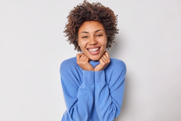 Mujer sonríe broady mantiene la mano en el cuello del suéter azul feliz de escuchar cumplidos disfruta de la vida expresa emociones positivas aisladas en blanco