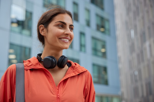 Mujer sonríe felizmente regresa del entrenamiento físico lleva auriculares estéreo alrededor del cuello vestido con rompevientos está afuera