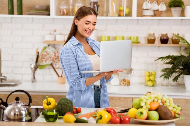 Una mujer sonríe mientras mira una computadora portátil en su cocina rodeada de productos frescos