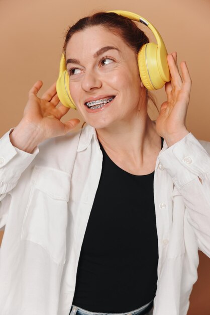 Foto mujer sonriendo con aparatos ortopédicos en los dientes y escuchando música en auriculares sobre un fondo beige