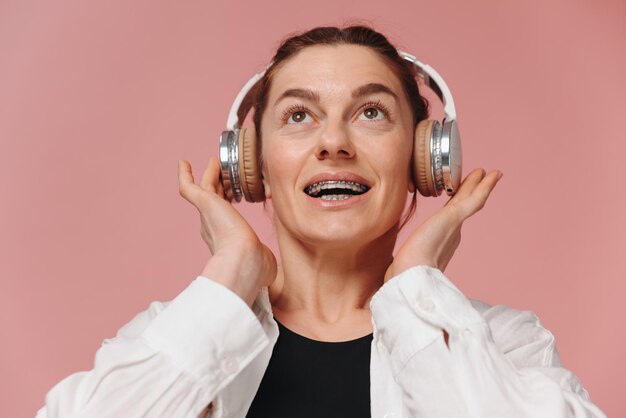 Foto mujer sonriendo con aparatos ortopédicos en los dientes y escuchando música en auriculares sobre un fondo rosa