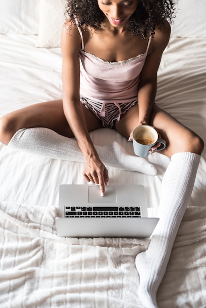 Mujer sonriendo bebiendo un café y trabajando con su computadora en la cama