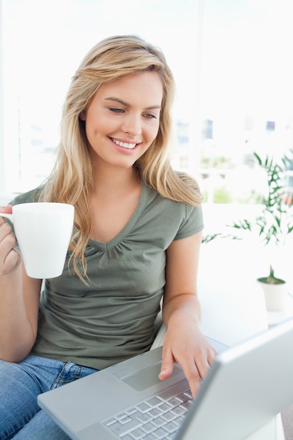 Mujer sonriendo mientras usa su computadora portátil y sostiene una taza en su mano libre