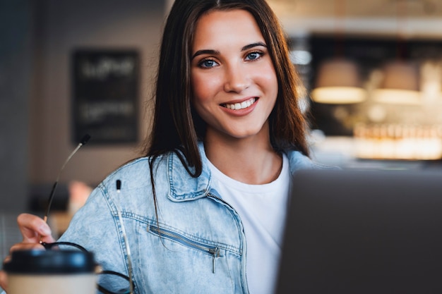 Mujer sonriendo, mirando a la cámara. Mujer de negocios atractiva se sienta a la mesa frente a conversaciones de portátil en el teléfono móvil