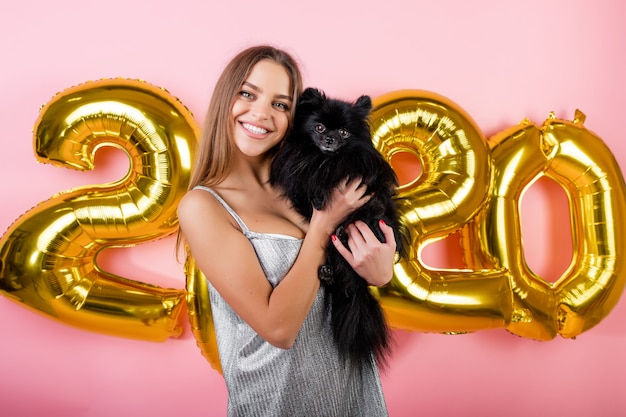 Mujer sonriendo con spitz japonés perro negro y globos de oro 2020 año nuevo aislados en rosa