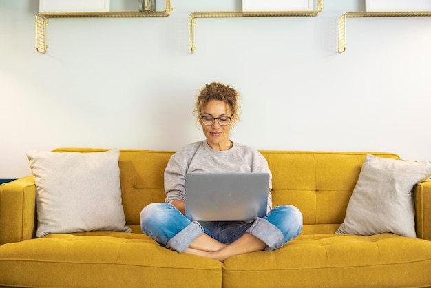 Una mujer sonriendo y usando una computadora portátil en casa sentada cómodamente en un sofá amarillo en la sala de estar Computadora portátil de personas trabajadoras inteligentes Navegando por la web Disfrutando de la tecnología y la conexión