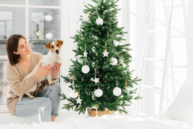 La mujer sonriente abraza a la mascota con amor tiene un estado de ánimo feliz posan juntos cerca de un hermoso árbol de Navidad decorado paredes blancas escalera y ventana alrededor pasan tiempo en casa vacaciones de invierno