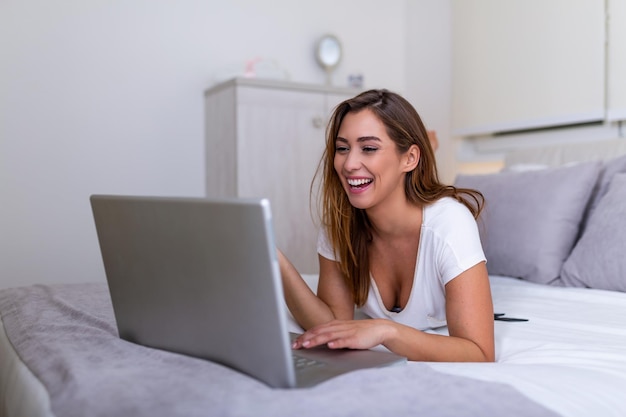 Mujer sonriente acostada en la cama frente a su computadora portátil con las piernas levantadas. Feliz hermosa mujer casual trabajando en una computadora portátil sentada en la cama en la casa. Freelance trabajando desde el concepto de hogar