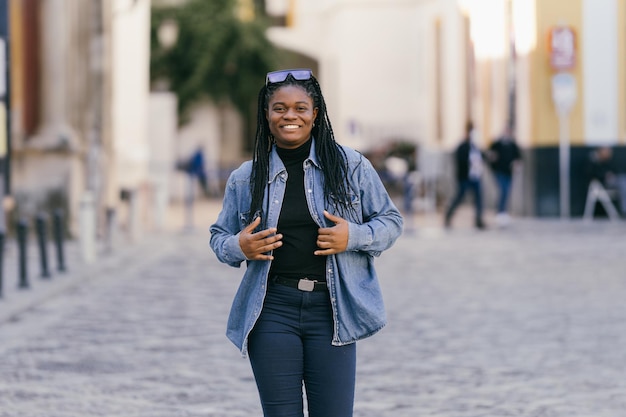 Mujer sonriente africana en ropa de mezclilla y gafas de sol en la cabeza caminando por la calle