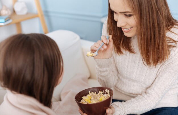 Mujer sonriente alegre dando desayuno a su hija mientras está sentada en el sofá