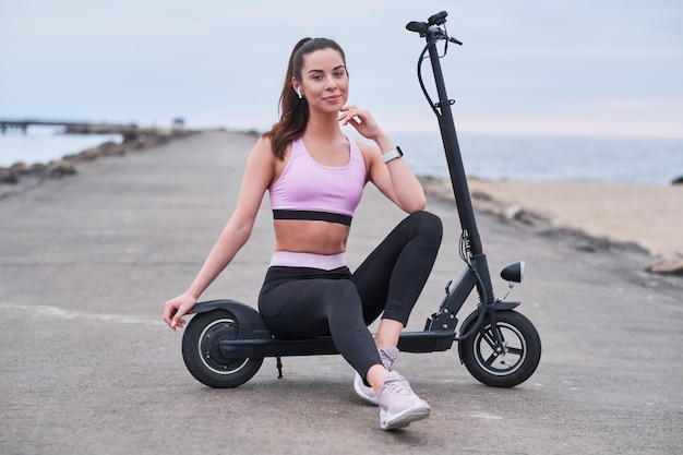 Mujer sonriente alegre se está enfriando en su nuevo scooter eléctrico en la playa en un día nublado.