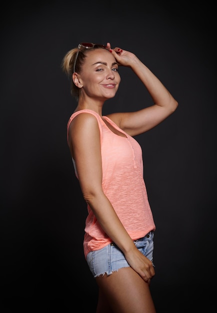 Mujer sonriente alegre con piel bronceada hermoso cuerpo musculoso en pantalones cortos de mezclilla mirando a través de sus gafas de sol rosas y mostrando la lengua a la cámara posando sobre el espacio de copia de fondo negro