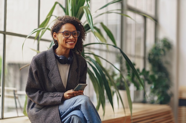 Mujer sonriente con anteojos hablando por teléfono con amigos mientras se sienta en el coworking cerca de las ventanas