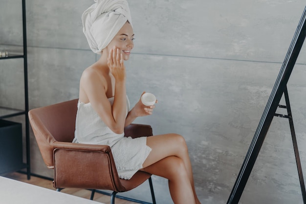 Mujer sonriente aplica toalla envuelta en crema facial en la cabeza cuida la piel Rutina de belleza