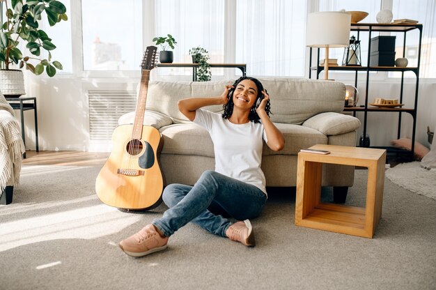 Mujer sonriente en auriculares en casa, guita. Señora bonita con instrumento musical relajarse en la habitación, amante de la música femenina descansando