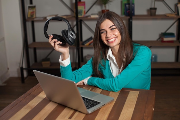 Una mujer sonriente con auriculares negros está sentada en la mesa con una laptop gris