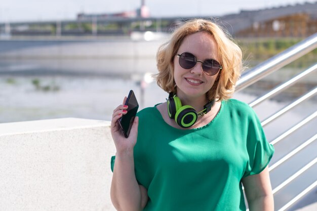 Una mujer sonriente con auriculares sosteniendo un teléfono en la mano escucha música en la calle