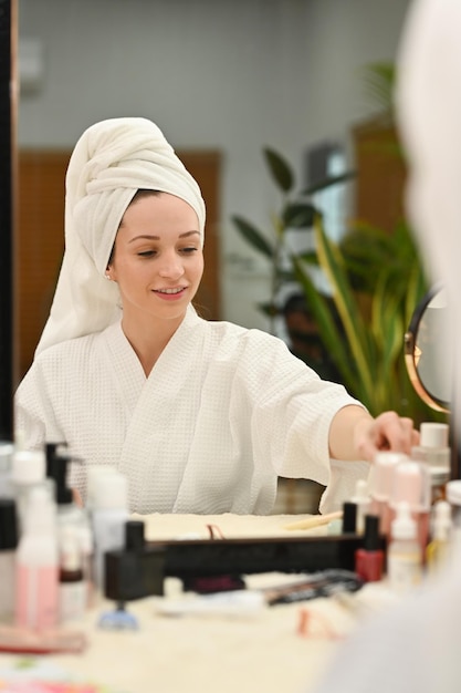 Mujer sonriente en bata de baño sentada frente al espejo haciendo rutina matutina después de la ducha Concepto de cuidado personal y tratamiento de belleza