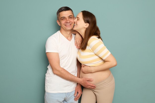Mujer sonriente besando a su marido en la mejilla embarazada pareja joven caucásica en ropa casual de pie aislada sobre un fondo verde claro amando a la familia esperando un bebé