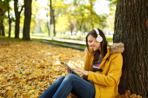 La mujer sonriente de cabello castaño con abrigo amarillo y jeans sentado y escuchando música debajo de un árbol con una tableta en sus manos y auriculares en el parque de la ciudad de otoño en un día caluroso. Otoño de hojas doradas.