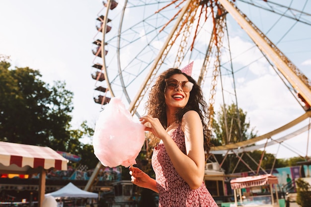 Mujer sonriente con cabello rizado oscuro con gafas de sol y gorra de cumpleaños de pie con algodón de azúcar en la mano y felizmente mirando a un lado mientras pasa tiempo en el parque de diversiones con la rueda de la fortuna en el fondo