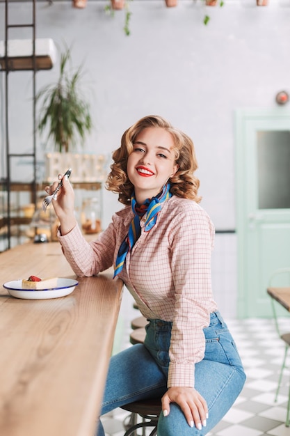 Mujer sonriente con camisa y jeans sentada en la barra del café con tenedor en la mano y pastel cerca y mirando alegremente a la cámara