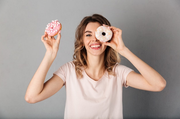 Mujer sonriente en camiseta divirtiéndose con donas y sobre gris