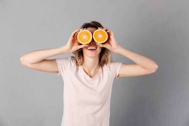 Mujer sonriente en camiseta divirtiéndose y sosteniendo naranjas en sus ojos sobre gris