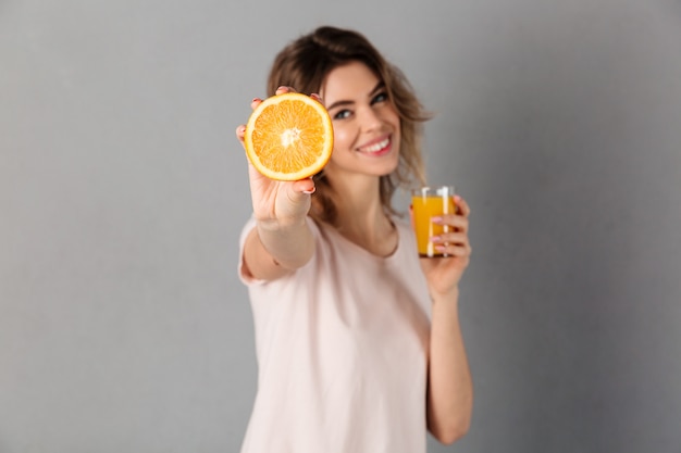 Mujer sonriente en la camiseta que muestra la naranja mientras que sostiene la taza de jugo sobre gris. Centrarse en naranja