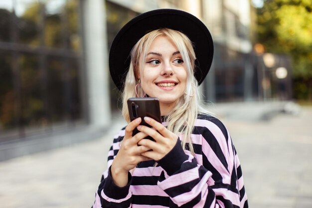 Mujer sonriente carismática joven en sombrero usando el teléfono al aire libre