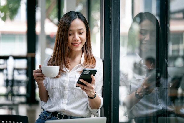 Mujer sonriente en casual de pie en la oficina Mujer de negocios con teléfono móvil y una taza de café en la mano relajándose durante el trabajo