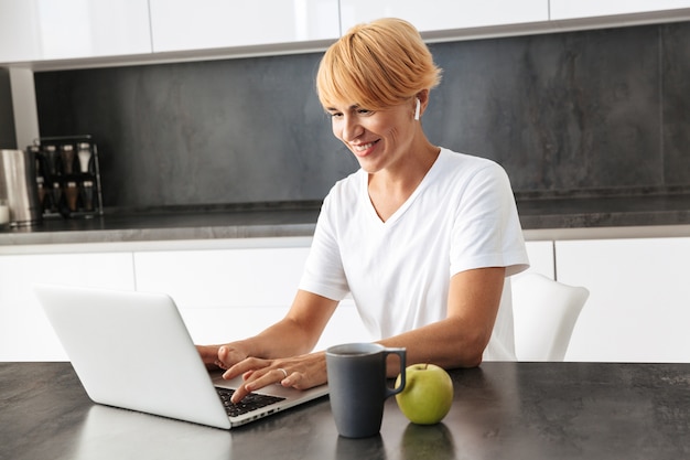 Mujer sonriente casualy vestida con ordenador portátil mientras está sentado en una cocina, con auriculares