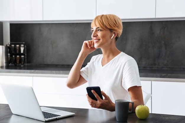 Mujer sonriente casualy vestida con ordenador portátil mientras está sentado en una cocina, con auriculares