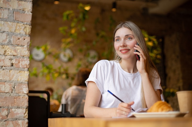 Mujer sonriente caucásica con una camiseta blanca mira hacia un lado mientras habla por teléfono y escribe en un papel con un croissant y café en un café en la mesa