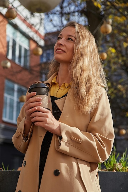 Mujer sonriente caucásica en gabardina beige sosteniendo una taza de café al aire libre