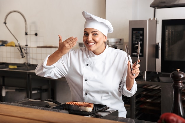 Mujer sonriente chef cocinero vistiendo uniforme cocinando delicioso filete de salmón de pie en la cocina