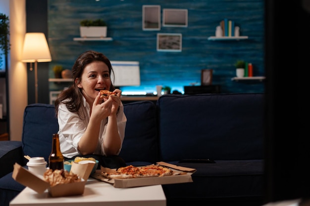 Mujer sonriente comiendo una rebanada de entrega de pizza caliente sentada en el sofá mirando el programa familiar en la televisión en la sala de estar. Persona feliz disfrutando de la cena de televisión para llevar en la mesa con comida rápida para llevar.