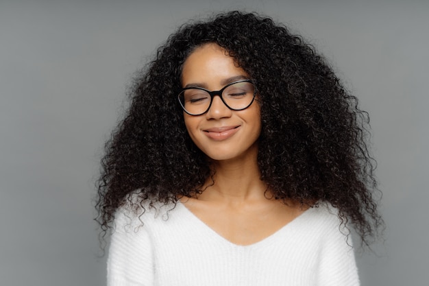 Mujer sonriente complacida con el cabello tupido y crujiente, mantiene los ojos cerrados, sonríe suavemente, usa lentes ópticos