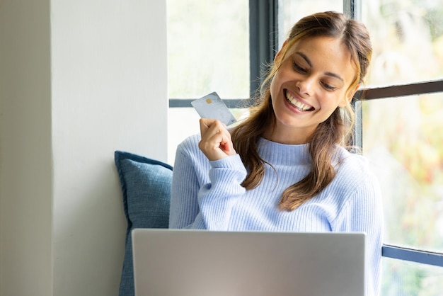 Mujer sonriente comprando en línea con tarjeta de crédito en casa cerca de la ventana