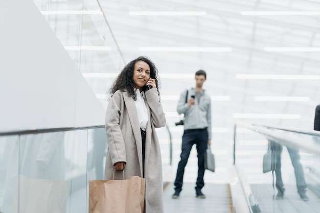 Mujer sonriente con compras de pie en la escalera mecánica en el centro comercial