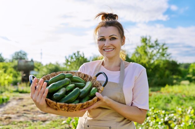 Mujer sonriente con cosecha de pepinos en la granja, jardinera femenina en el soleado huerto de verano con cesta de pepinos. Aficiones, jardinería, cultivo de hortalizas orgánicas.