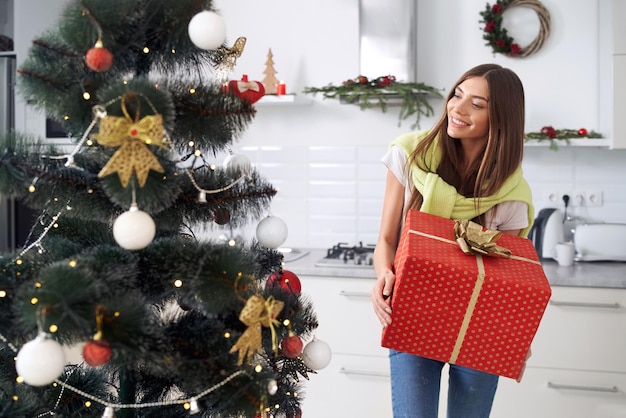 Mujer sonriente disfrutando del árbol de Navidad y sosteniendo regalo rojo