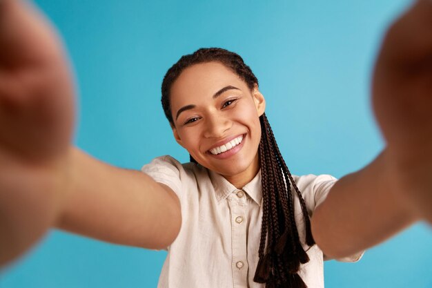 Mujer sonriente con dreadlocks negros tiene expresión de cara feliz, estira el brazo para hacer selfie, expresa emociones sinceras, foto de punto de vista. Disparo de estudio interior aislado sobre fondo azul.