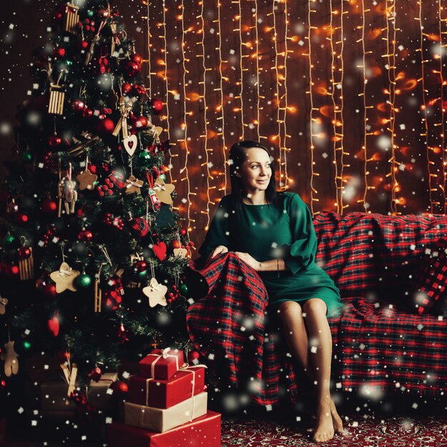 Foto mujer sonriente en elegante vestido verde en el sofá en el árbol de navidad.