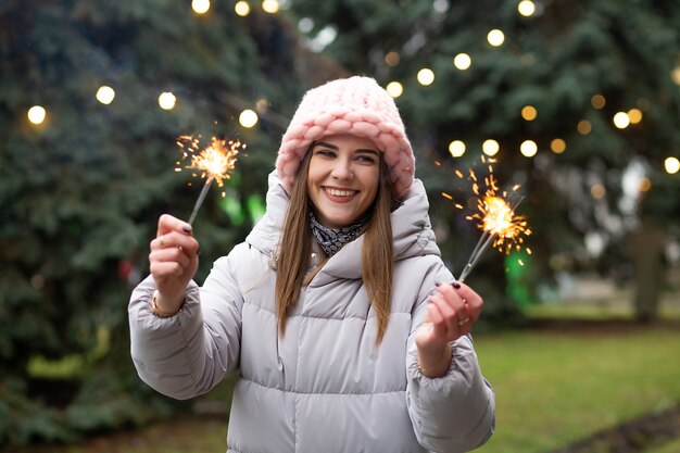 Mujer sonriente emocionada divirtiéndose con bengalas cerca del árbol de año nuevo