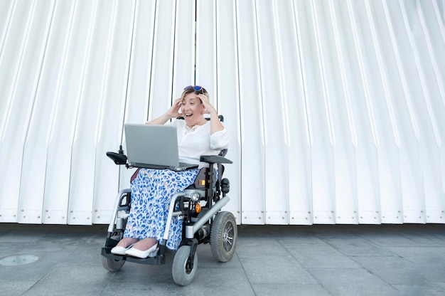 Una mujer sonriente y emocionada en silla de ruedas tomándose de la mano en la cabeza haciendo una videollamada usando la laptop al aire libre en un espacio de copia de fondo blanco