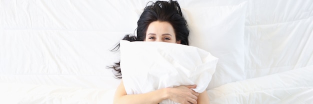 Mujer sonriente se encuentra en la cama blanca y abraza la almohada para dormir sano y luchar contra el insomnio