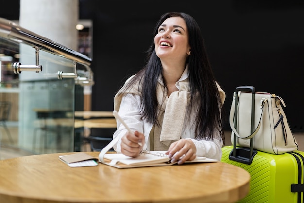 Mujer sonriente escribiendo tomando notas en el diario sentado a la mesa en la terminal del aeropuerto internacional