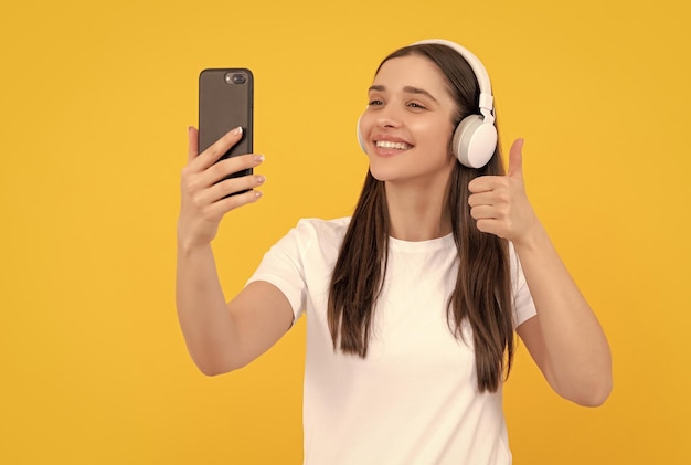 Mujer sonriente escucha música en auriculares con teléfono inteligente muestra el pulgar hacia arriba en selfie de fondo amarillo