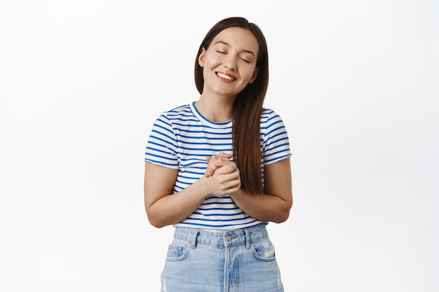 Mujer sonriente esperanzada soñando con algo, tomarse de las manos juntas, con los ojos cerrados y soñando despierto, pensando en cosas hermosas, de pie en una camiseta a rayas contra la pared blanca.